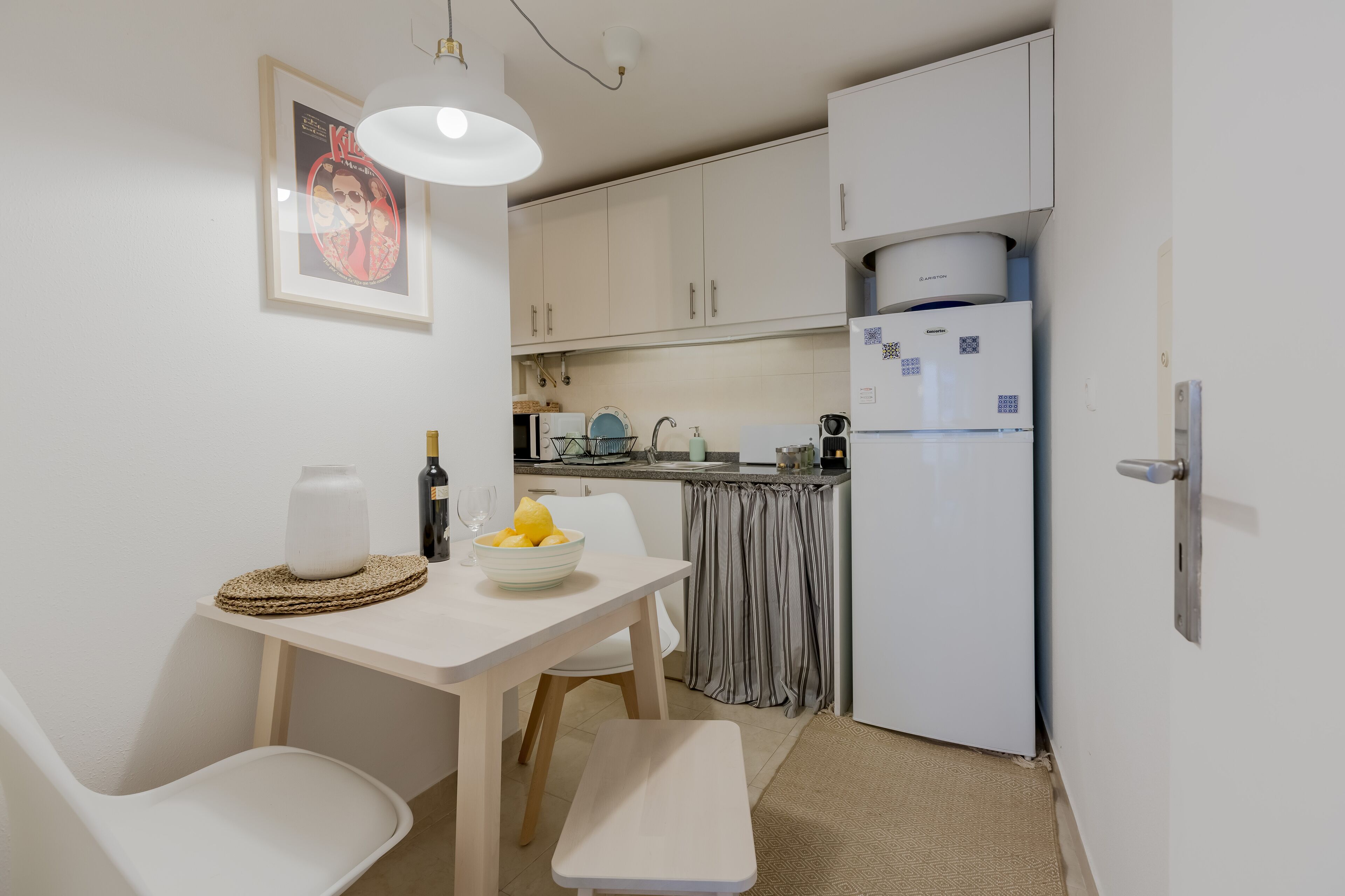 Une cuisine compacte avec des meubles blancs, un réfrigérateur blanc, une table à manger avec des chaises et un luminaire suspendu.