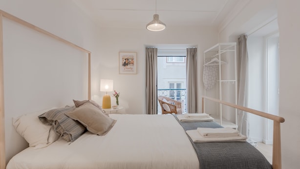 A bedroom with a large bed, a bedside table with a lamp, a chair, a balcony with a view, and a wardrobe. - Sunny Bairro Alto / Chiado Apartment, By TimeCooler (Lisbon)