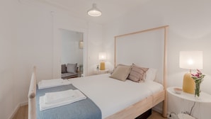 A neatly made bed with white linens, a blue throw blanket, and a wooden headboard. There is a small bedside table with a lamp and a vase of flowers. The room has white walls and a wooden floor. - Sunny Bairro Alto / Chiado Apartment, By TimeCooler (Lisbon)