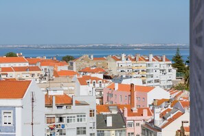 A coastal town with terracotta-roofed buildings, a clear view of the sea, and a distant cityscape. - Renovated Sunny Apartment with Balcony, By TimeCooler (Lisbon)