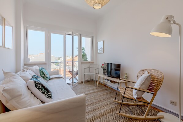 A modern bedroom with a bed, a TV, a rocking chair, and a view of the city through large windows. - Renovated Sunny Apartment with Balcony, By TimeCooler (Lisbon)