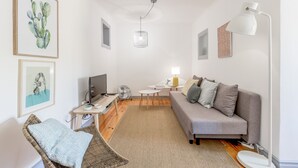 Ein modernes Wohnzimmer mit grauem Sofa, einem Korbstuhl, einem Flachbildfernseher, einem kleinen Tisch und einer Deckenleuchte.