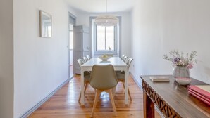 A dining area with a white table, grey chairs, a wooden floor, a hanging light fixture, and a vase with flowers on a wooden side table.