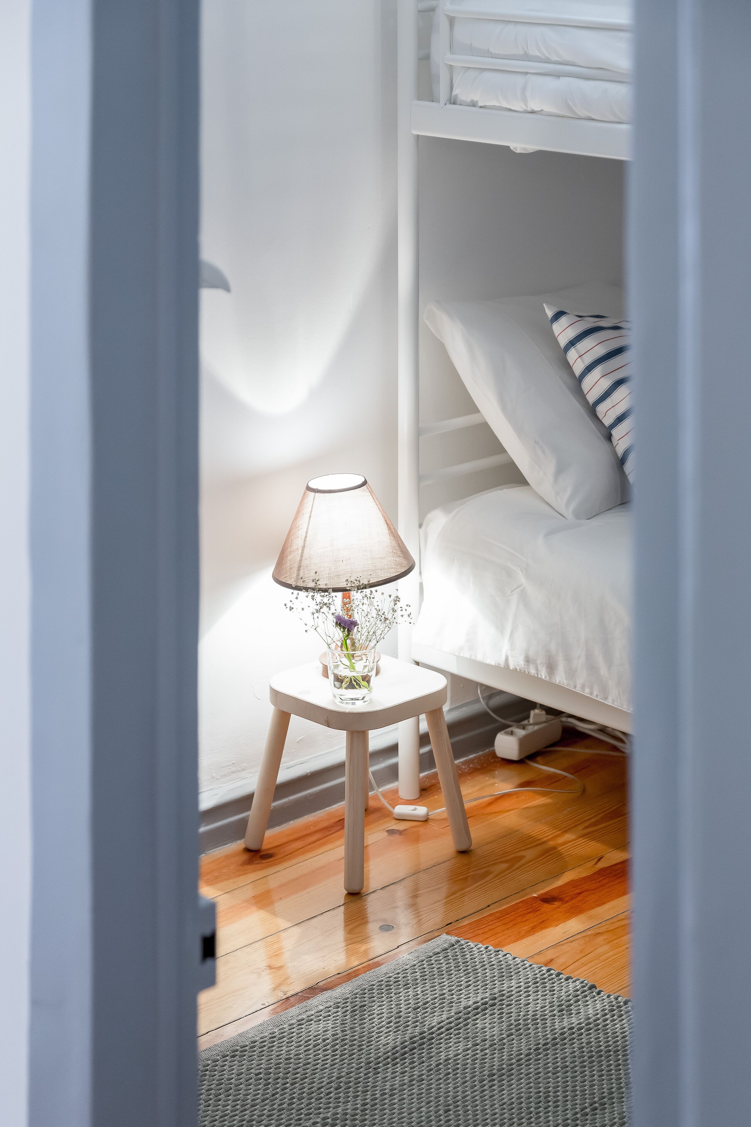 A room with a bunk bed, a small bedside table with a lamp, and a wooden floor.