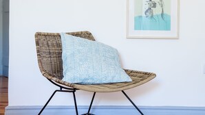A wicker chair with a patterned cushion, a framed cactus artwork, and a framed portrait on a white wall.