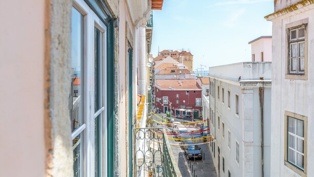 A narrow street with buildings on either side, a visible balcony with a railing, and a car parked on the street.