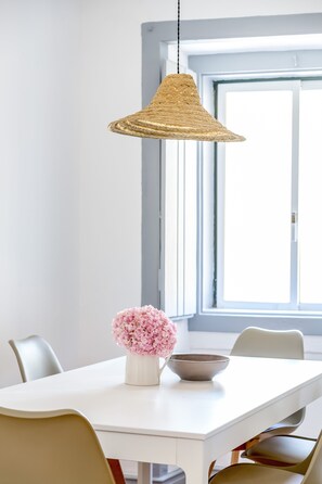A dining table with a white vase of pink flowers and a bowl, a woven pendant light, and chairs with a mix of colors. - Alfama, Bright Spacious W/ Terrace Apartment, By TimeCooler (Lisbon)