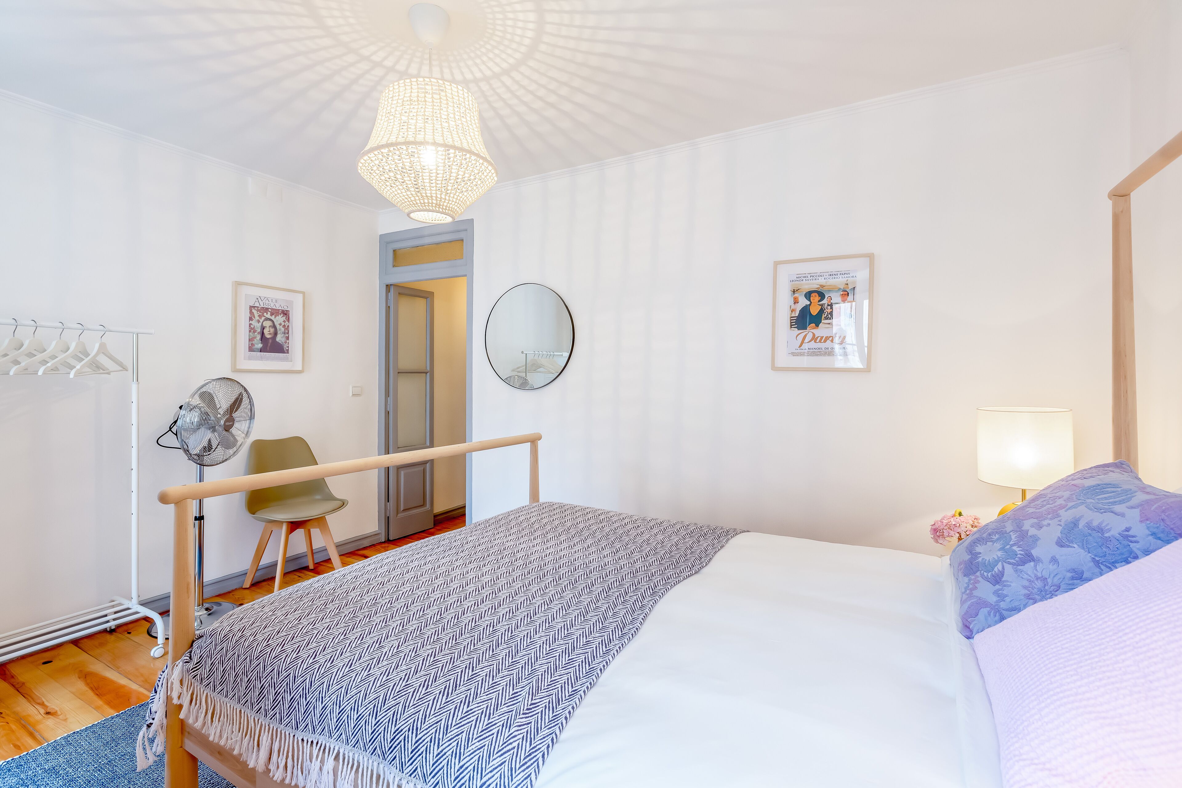 Une chambre à coucher comprenant un lit, une table de chevet avec une lampe, une chaise, un ventilateur et une porte donnant sur une autre pièce.