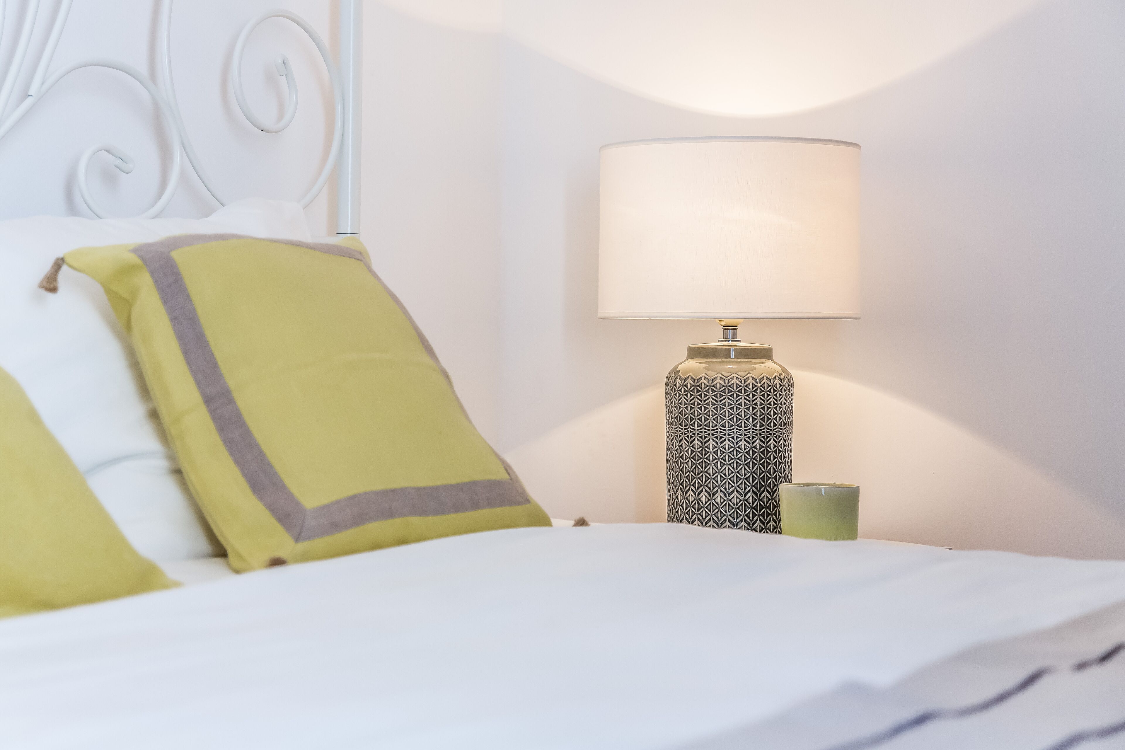 Ein ordentlich bezogenes Bett mit weißen und gelben Kissen, einer gemusterten Lampe und einem grünen Kerzenleuchter auf einem Nachttisch.