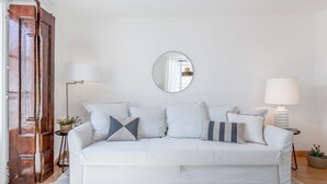 En vit soffa med flera kuddar, en rund spegel på väggen, en golvlampa och en dörr av trä.