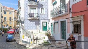 A narrow cobblestone street with buildings on either side, a woman standing by a railing, and a car parked on the left. - Typical Alfama Apartment, By TimeCooler (Lisbon)