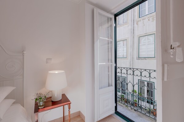 Un dormitorio con cama, una mesita de noche con lámpara y vistas a un balcón con plantas en macetas.