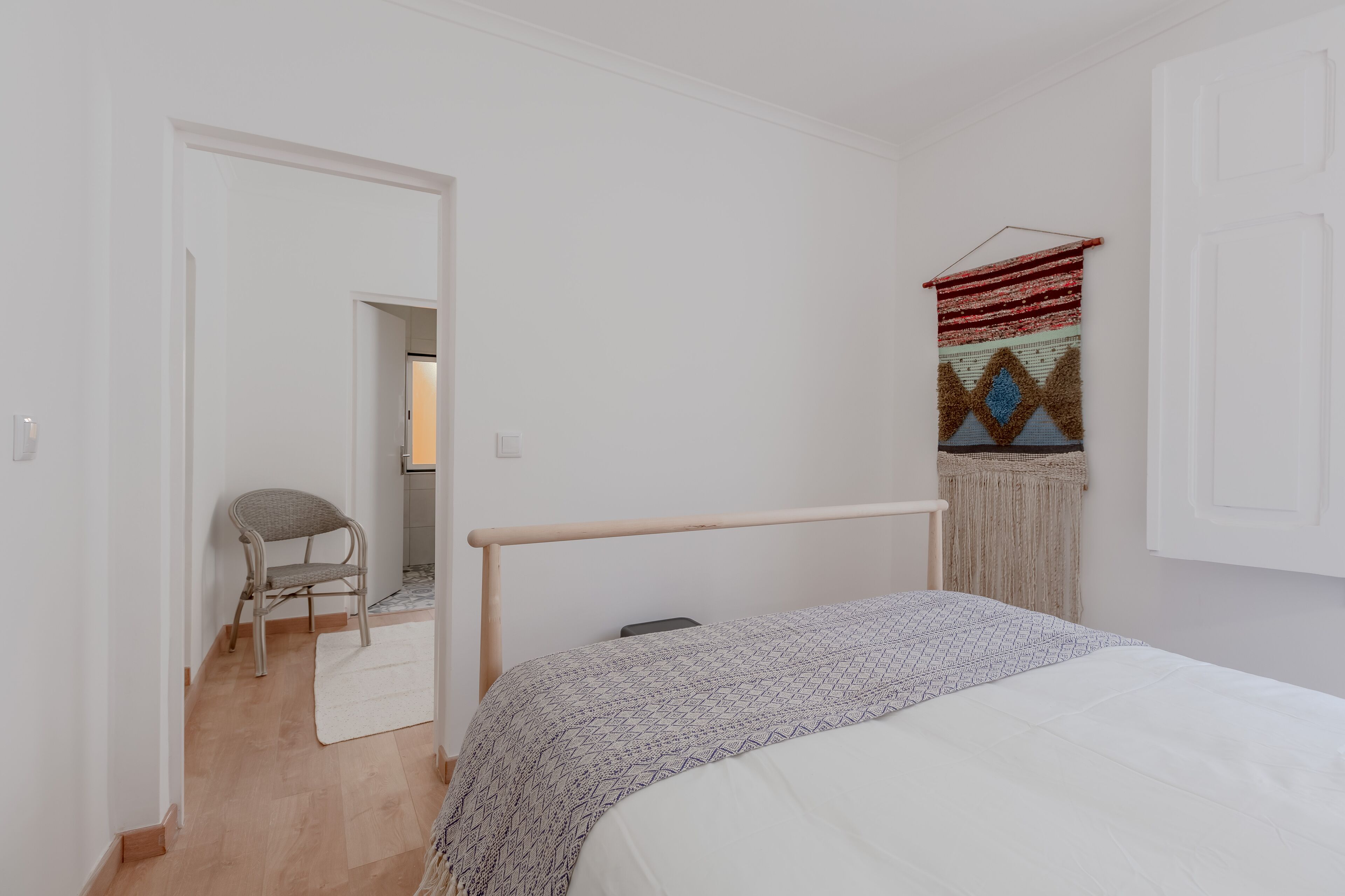 A bedroom with a bed, a chair, a hanging tapestry, and a door leading to another room.