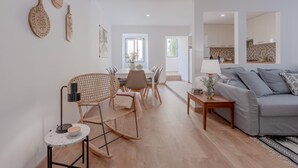 A modern living room with a sofa, a dining table with chairs, and a kitchen visible through an open doorway. - Bright & Spacious W/ Orange Tree Patio Apartment by TimeCooler (Lisbon)