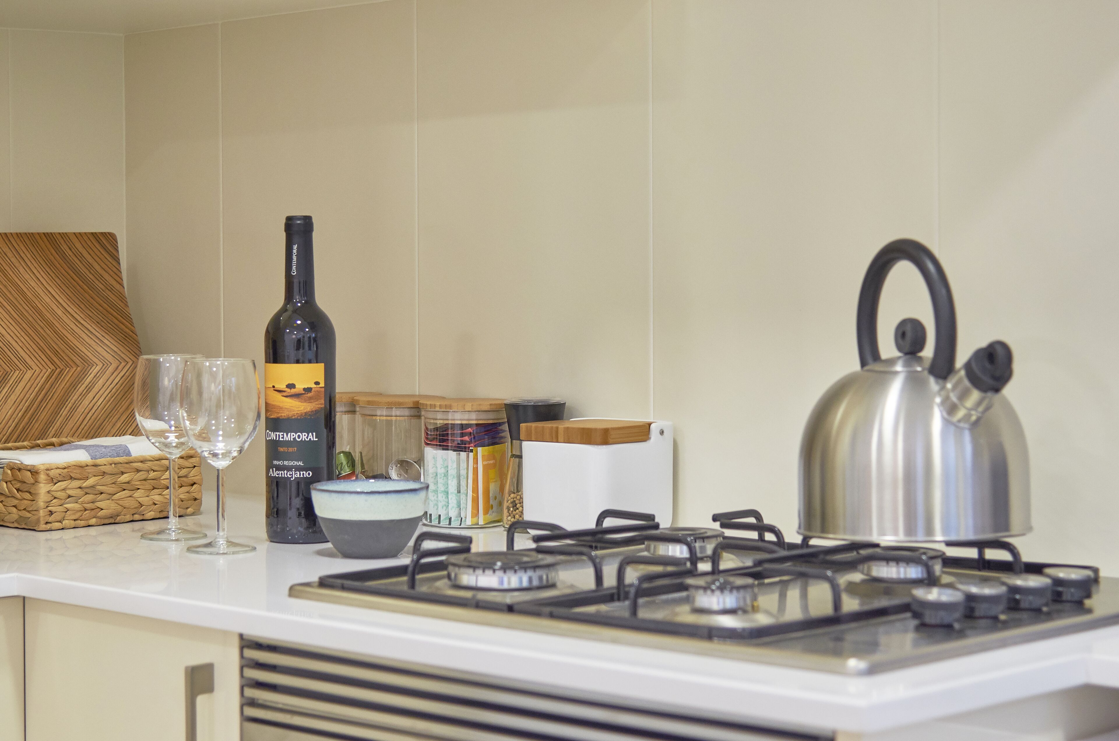 A kitchen counter with a bottle of wine, two glasses, a kettle, and a basket.