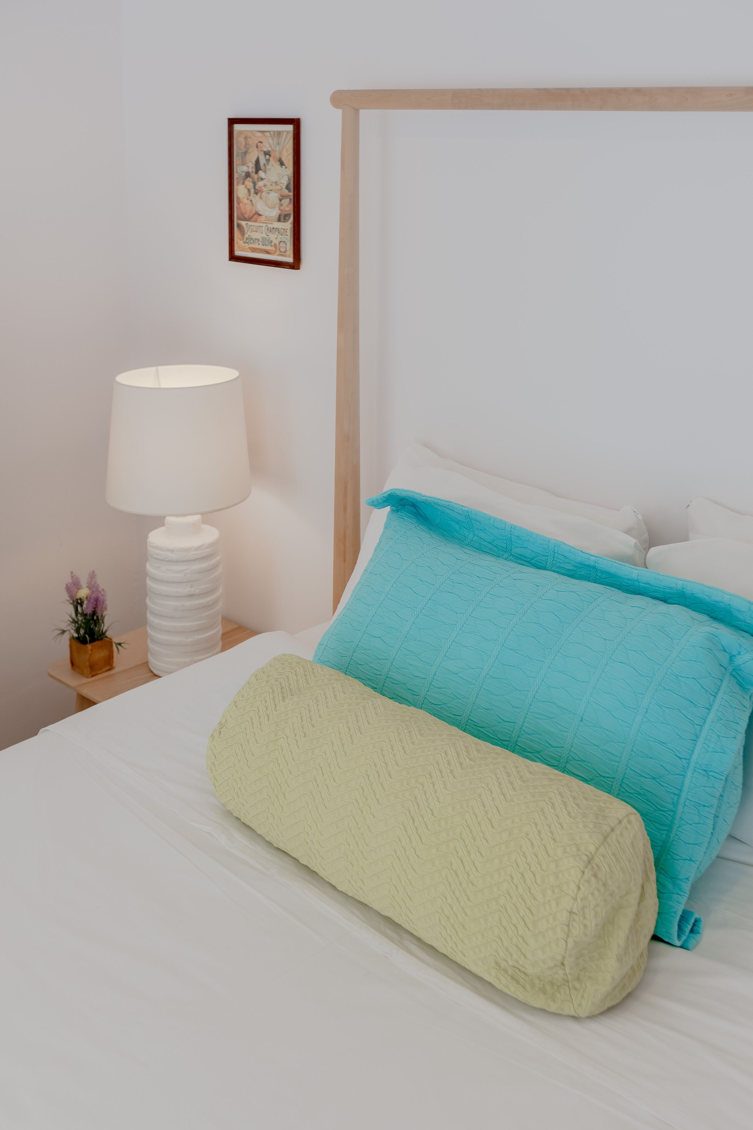 A neatly made bed with a white and teal quilt, a white lamp on a wooden nightstand, and a small vase with flowers.