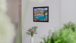 A modern interior with a white wall, a framed painting of a coastal scene, a white console table with a vase of flowers, and a potted plant. - Sunny Apartment with Patio, By TimeCooler (Lisbon)