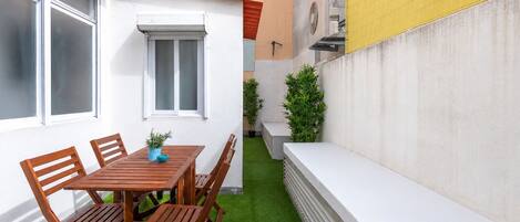 Une petite terrasse avec une table et des chaises en bois, un mur blanc et une pelouse artificielle verte.