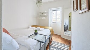 A bedroom with a bed, a bedside table with a plant, a hanging light fixture, a window, and a door leading to another room. - Alfama Light-Filled Apartment, By TimeCooler (Lisbon)