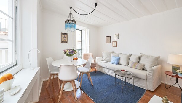 A bright living room with a white sofa, a dining table with chairs, and a blue rug. - Alfama Light-Filled Apartment, By TimeCooler (Lisbon)