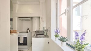 A compact kitchen with white cabinetry, a stove, a sink, and a window with potted plants. - Alfama Light-Filled Apartment, By TimeCooler (Lisbon)