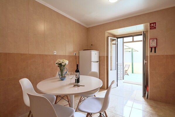 Un comedor con una mesa redonda, sillas, un florero con flores, una botella de vino y un refrigerador.