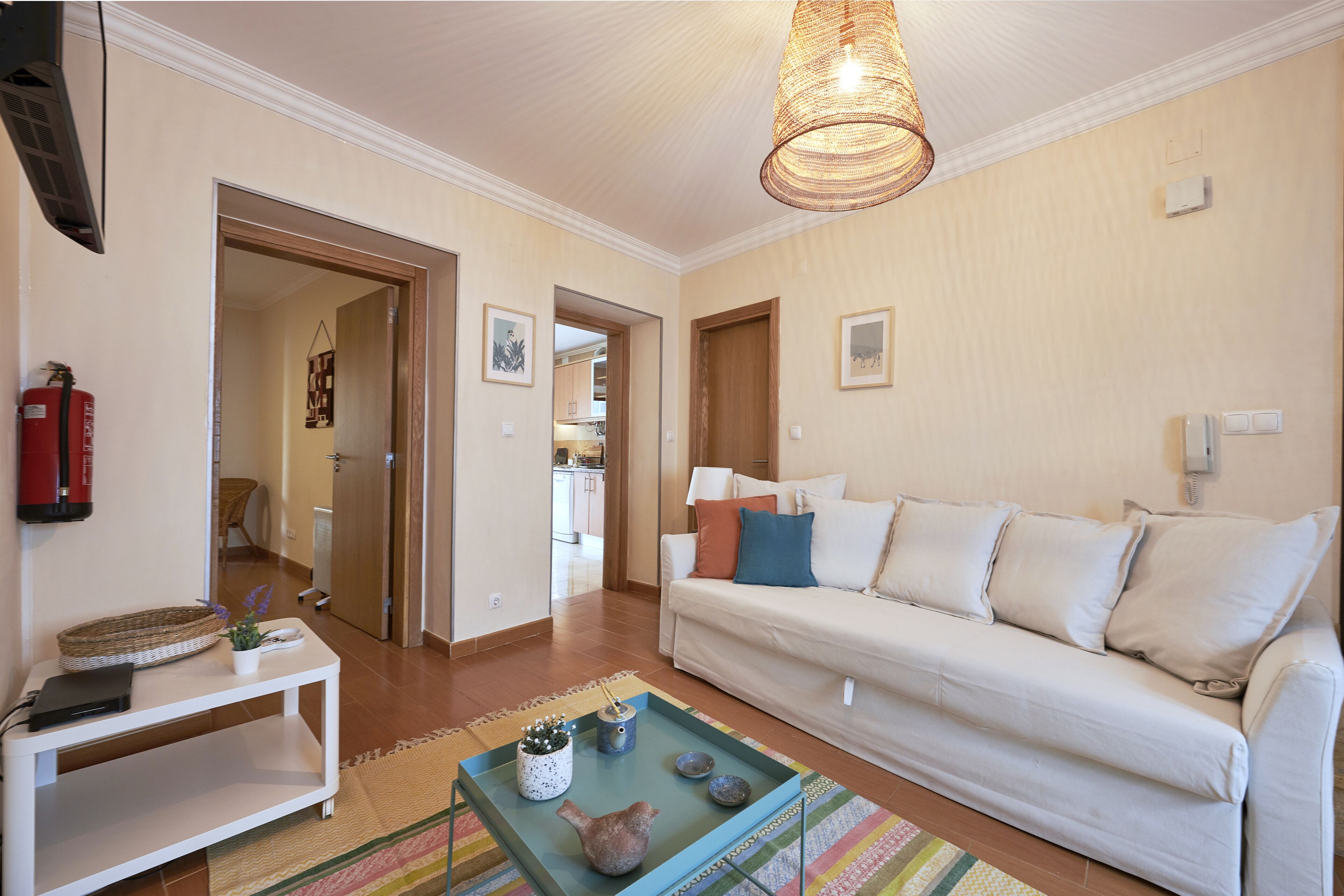 A living room with a white sofa, a coffee table with decorative items, a hanging light fixture, and a door leading to another room.