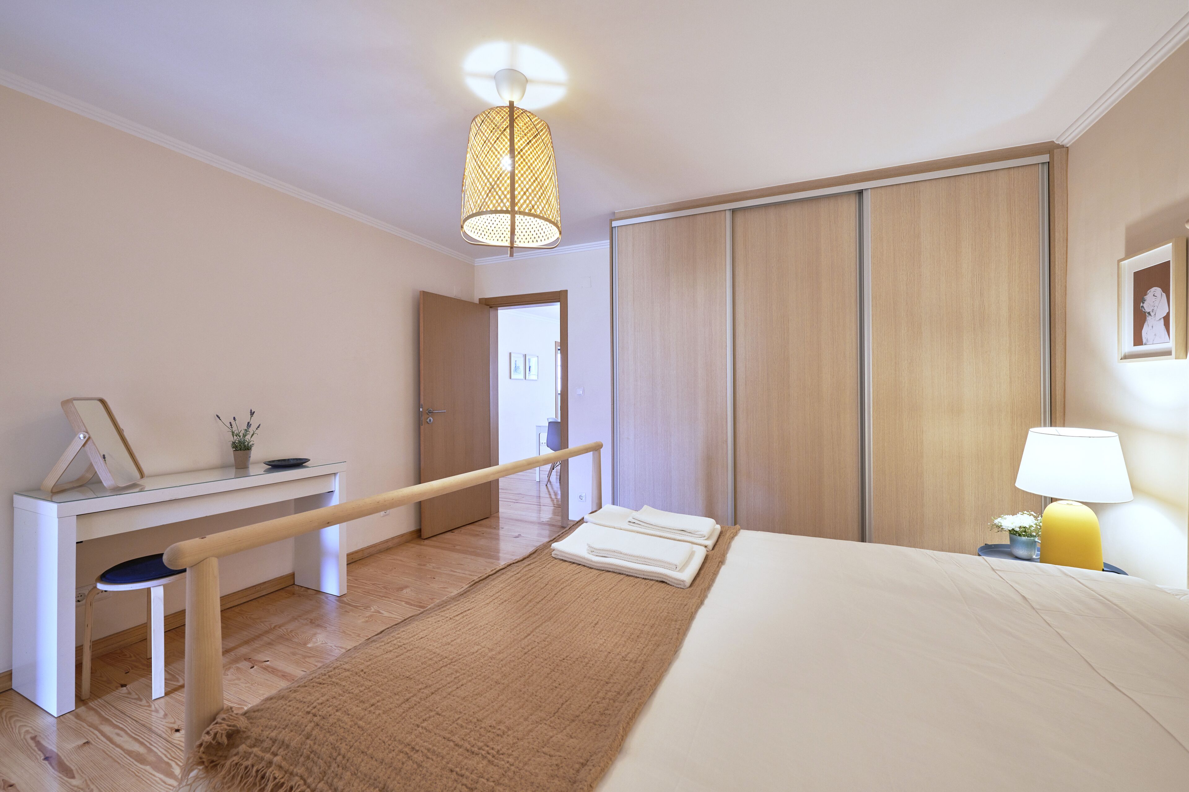 A bedroom with a bed, a desk, a chair, a bedside table with a lamp, and a wardrobe.