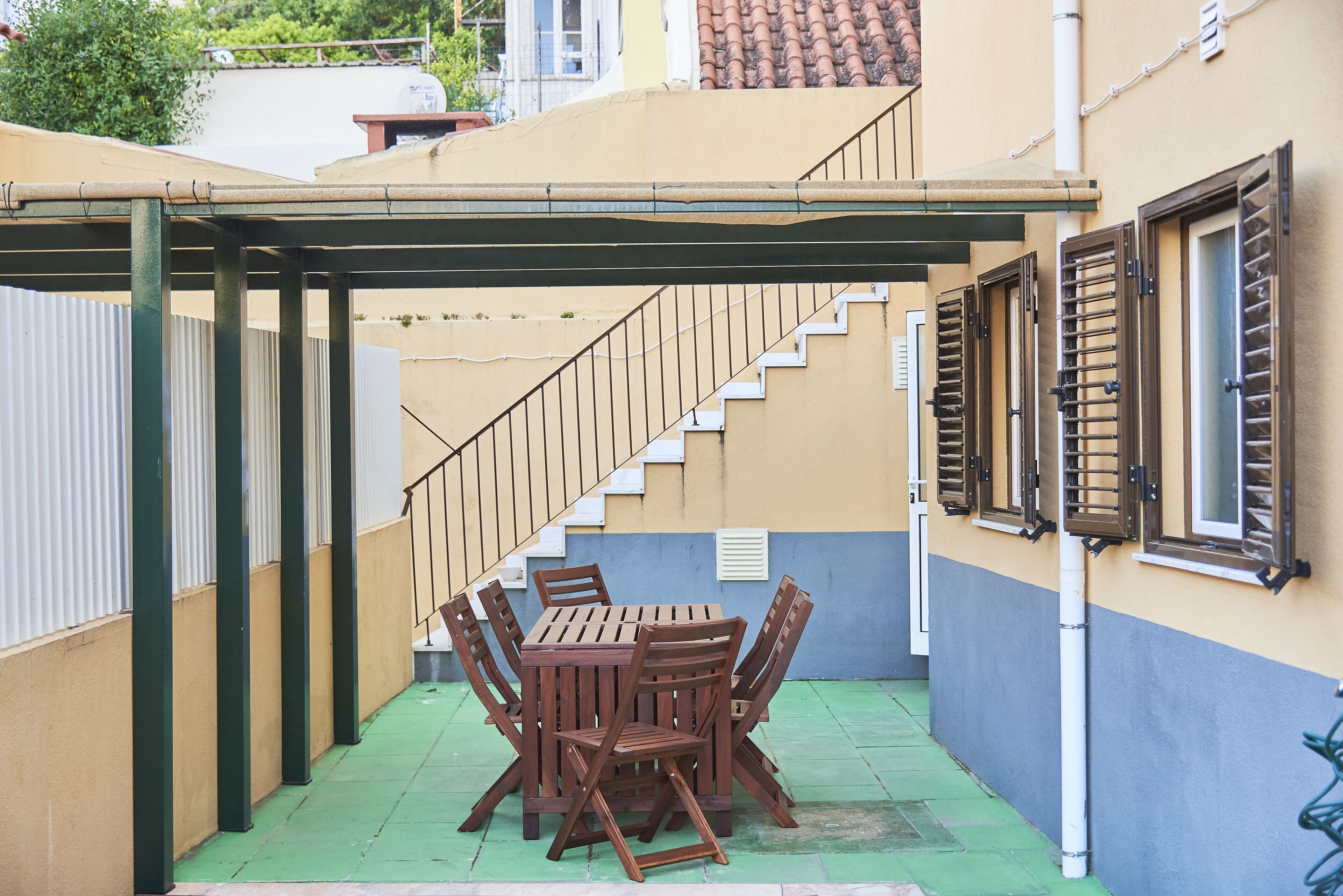 A covered outdoor patio with a wooden table and chairs, a staircase, and a building with shutters.
