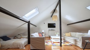 A loft-style bedroom with a sloped ceiling, a skylight, a flat-screen TV, a sofa, a coffee table, and a patterned rug.