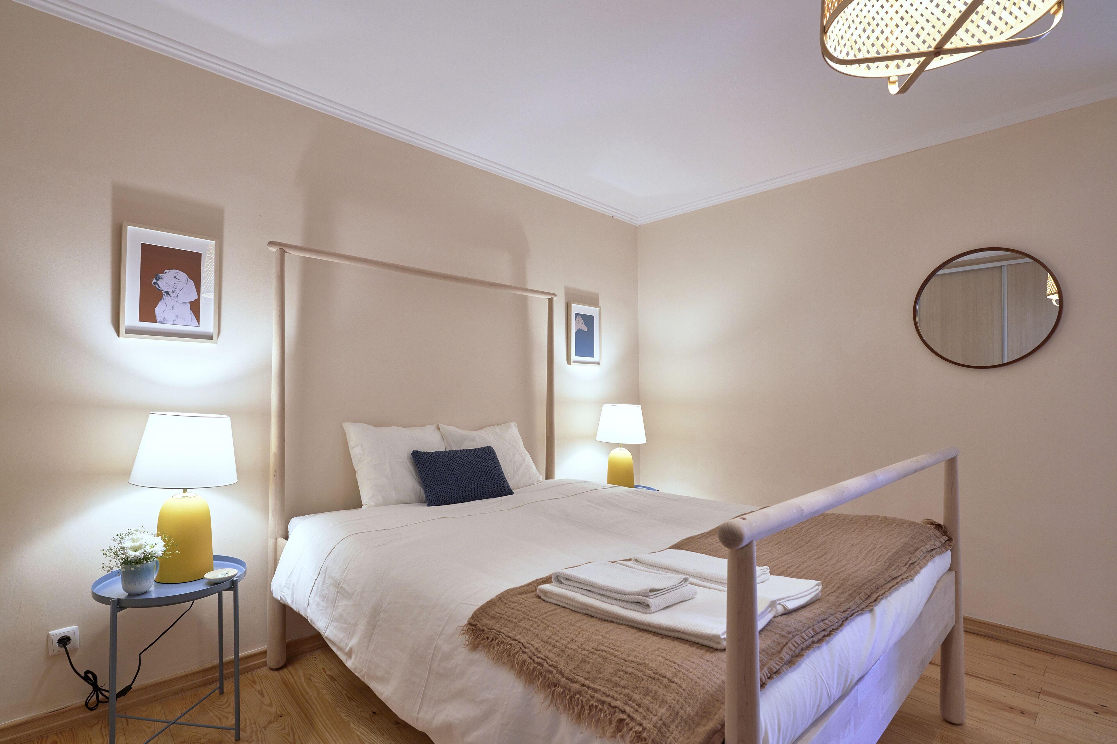 A neatly arranged bedroom with a bed, bedside lamps, a small table with a lamp, and framed pictures on the wall.