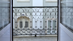 A view from a balcony with a metal railing, a building with two windows, and a door leading to a wooden floor.