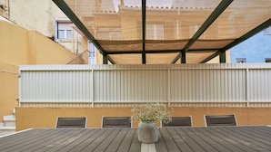 A covered outdoor dining area with a table and chairs, a potted plant, and a building in the background.