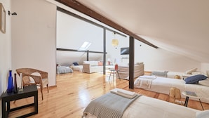 A spacious loft bedroom with a high ceiling, wooden beams, a large window, and a cozy seating area with a sofa and chairs.