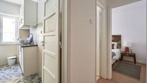 Eine kompakte Küche mit weißen Schränken, einem Spülbecken und einem gemusterten Boden. Durch einen offenen Durchgang ist ein Schlafzimmer mit Bett, Nachttisch und Lampe zu sehen.