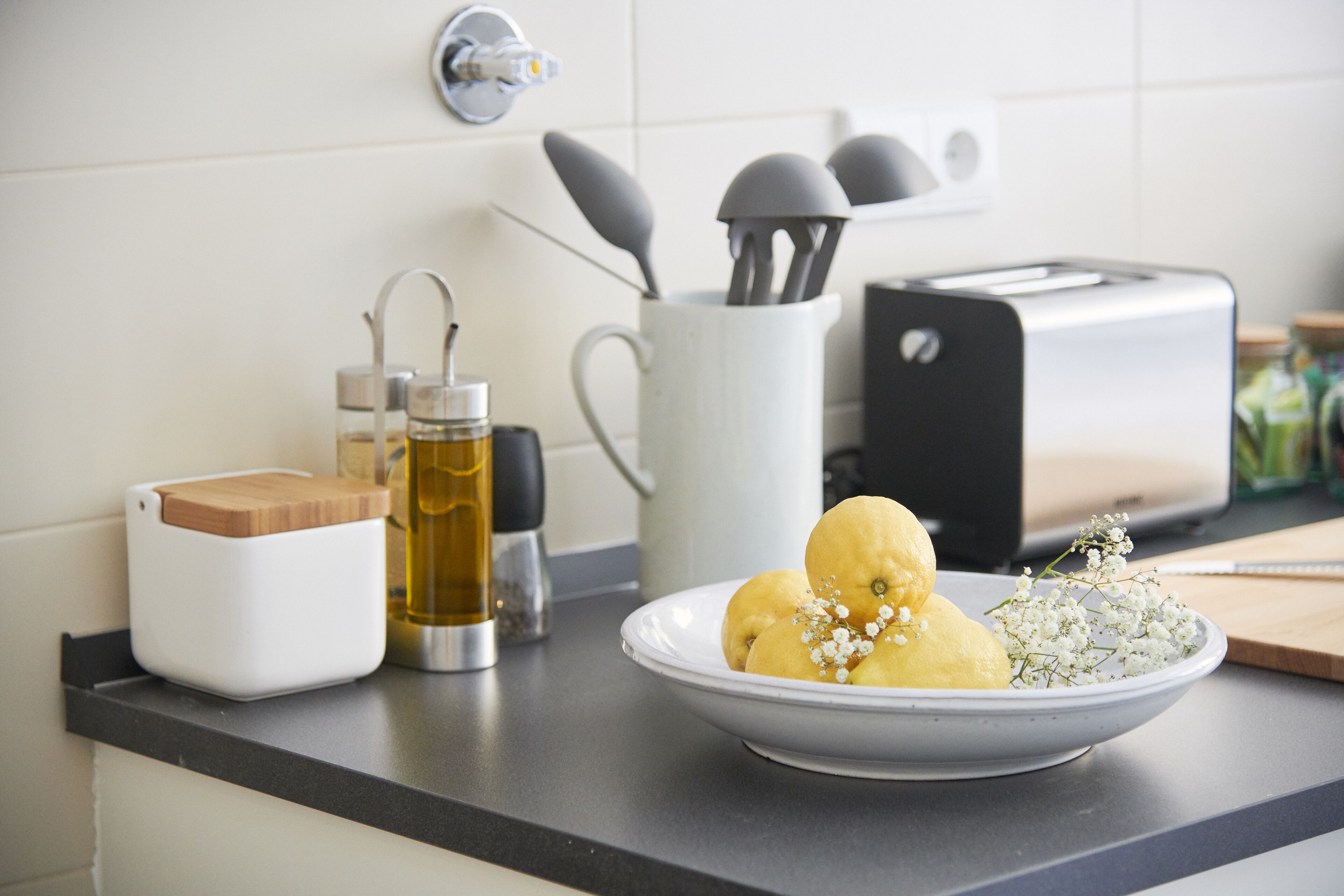 Una encimera de cocina con una taza blanca, una botella de aceite, una tabla de cortar y un plato de frutas amarillas.