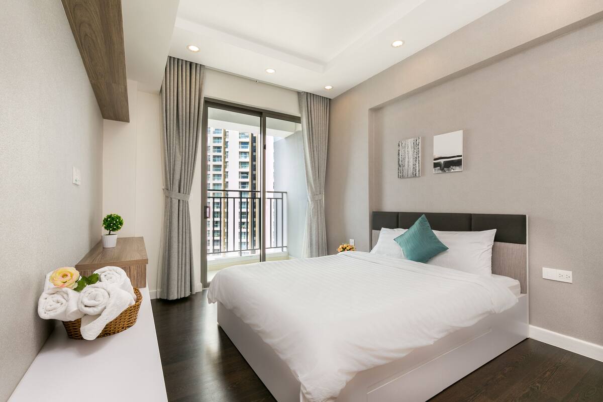 Ein modernes Hotelzimmer mit einem großen Bett, einem kleinen Beistelltisch mit Pflanze und Blick auf die Stadt vom Balkon aus.