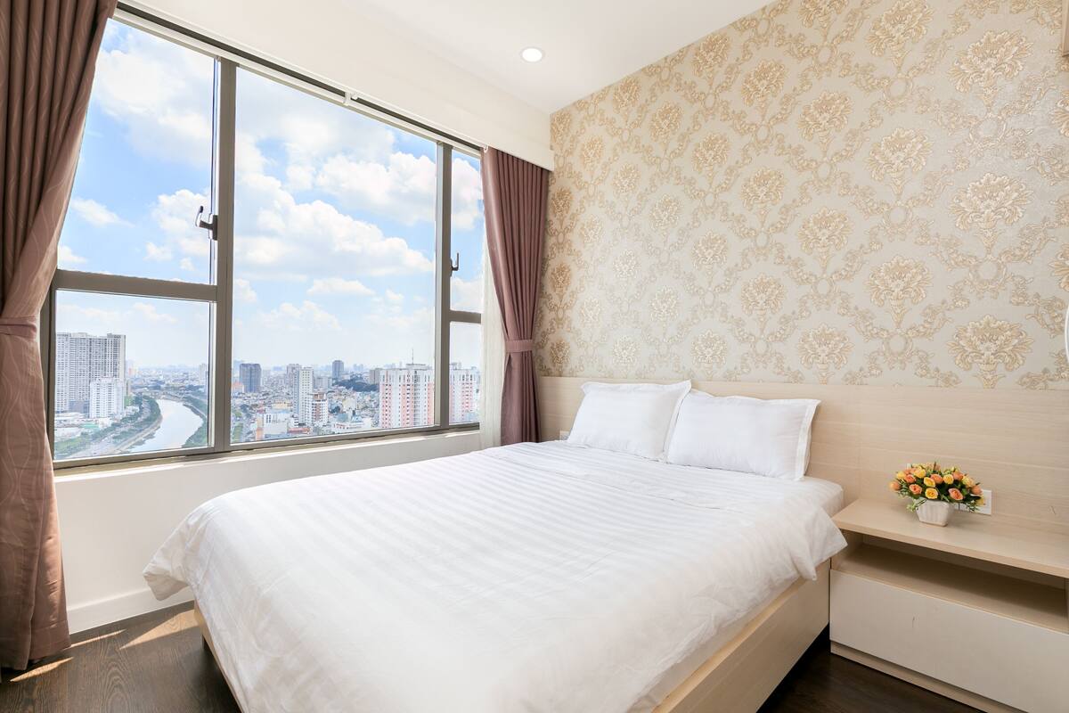 Ein Hotelzimmer mit großem Fenster, einem Bett, einem Nachttisch mit Blumenvase und floralem Wandpapier.