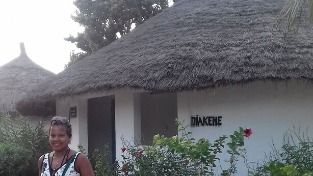 A person standing in front of a building with a thatched roof and a sign reading "DIAKENE". - Kounakito - Meaning 'welcome' (Oussouye)