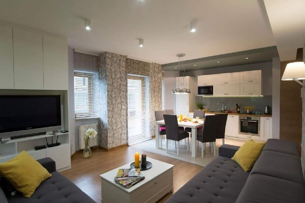 A modern living room with a flat-screen TV, grey sofas, and a dining area with a table and chairs.