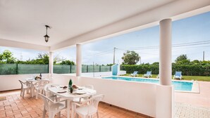 Un patio couvert avec des chaises blanches et une table, une piscine et une vue sur la végétation environnante.