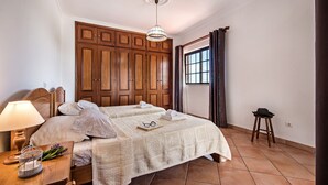 Une chambre avec une grande armoire en bois, un lit recouvert d’une courtepointe blanche, une table de chevet avec une lampe et une fenêtre avec des rideaux.