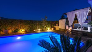 Piscina iluminada em azul à noite, cercada por plantas em vasos e uma pérgola com cortinas brancas.