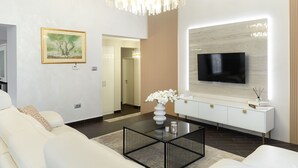 Uma sala de estar moderna com um sofá branco, uma mesa de centro de vidro, um candelabro, uma TV de tela plana presa a um painel de madeira e um quadro na parede.