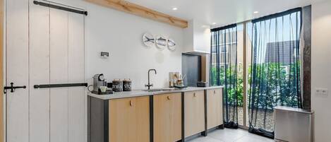 Cocina moderna con una isla central, un fregadero y un refrigerador. Se observan armarios de madera y un ventanal con cortinas translúcidas.