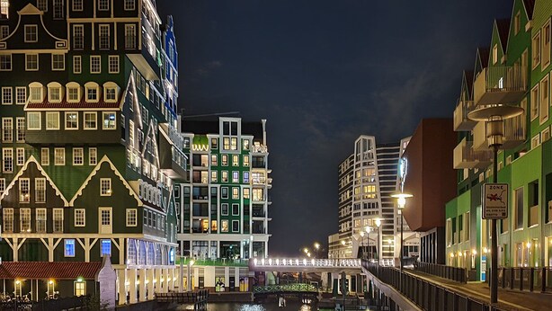 Ein Gebäude an einem Kanal, bei Nacht beleuchtet, mit mehreren Etagen, einer Brücke und einem Schild, das auf eine Zone hinweist.