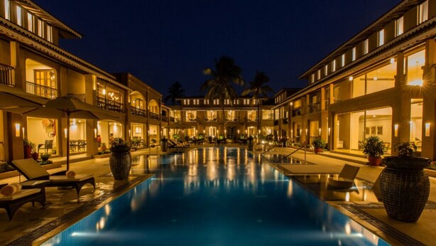 Zona de piscina hotelera con múltiples tumbonas y sombrillas, rodeada de edificios con balcones y grandes ventanales.