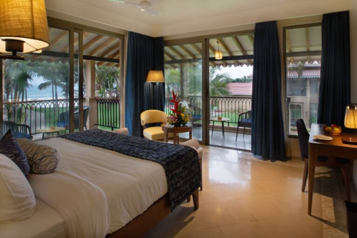 Quarto de hotel com uma cama grande, uma escrivaninha com abajur e uma varanda com vista para o exterior.