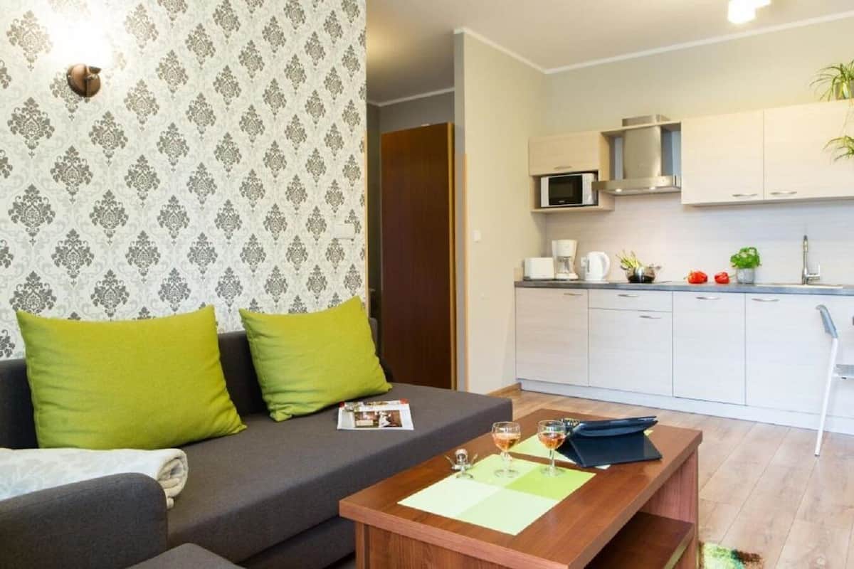 A modern living room with a grey sofa and green cushions, a wooden coffee table with glasses and a magazine, and a kitchen with white cabinets and a microwave.