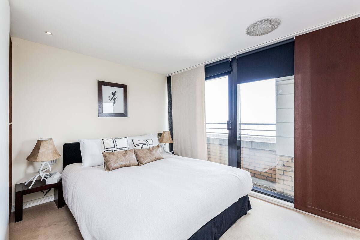 A modern hotel room with a large bed, a nightstand, a lamp, and a window with a view of the outside.